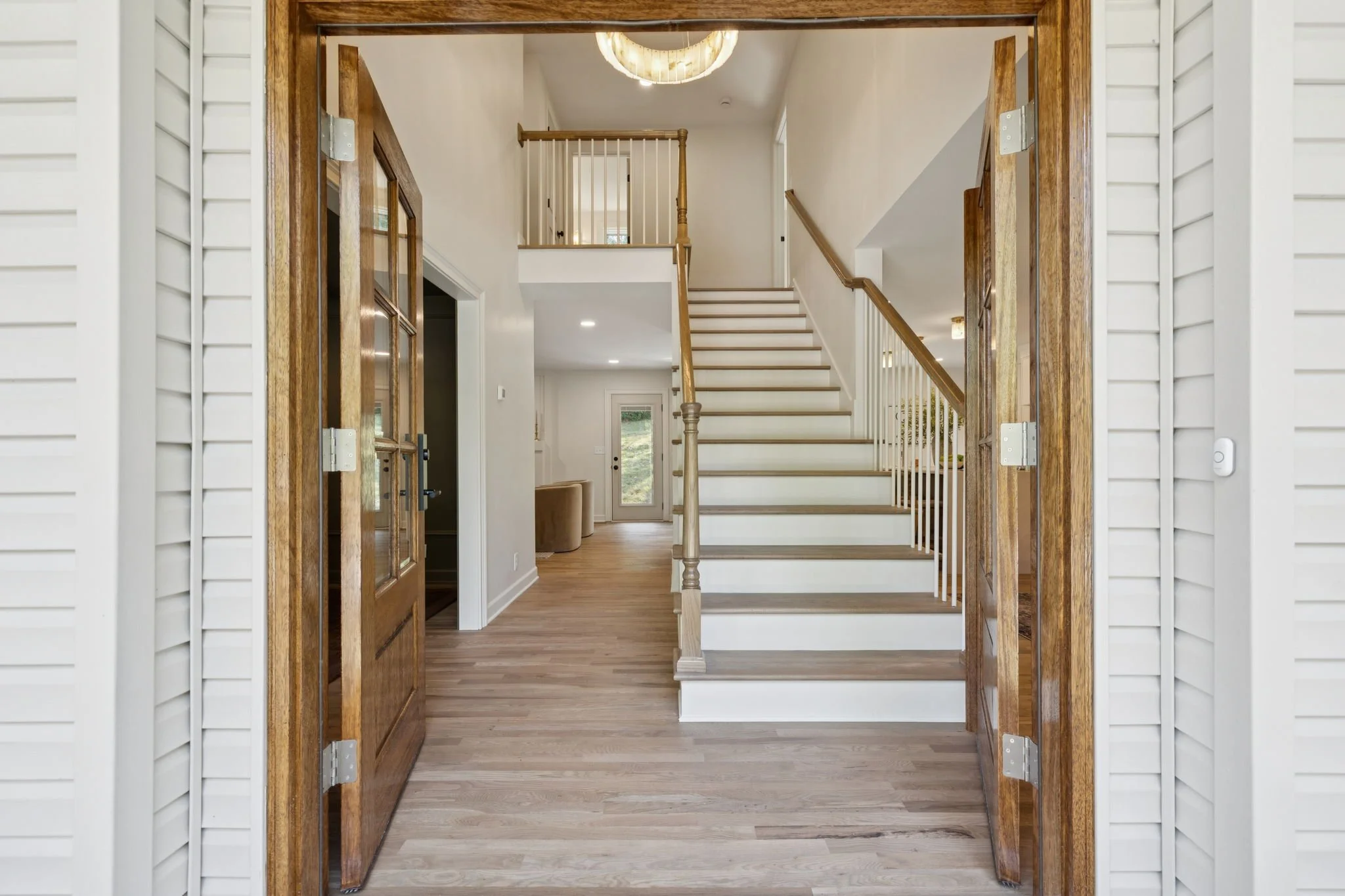 View through open front door into a house interior with a staircase, railing, doorways, hardwood flooring, and a ceiling light fixture.