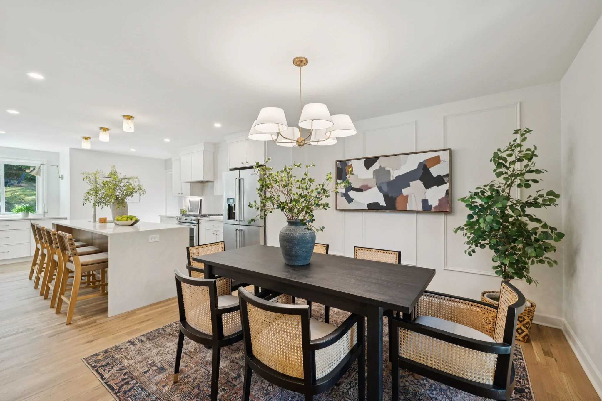 Open-concept kitchen and dining area with white walls, hardwood floors, modern lighting fixtures, framed abstract artwork, large potted plants, and a black dining table with chairs.