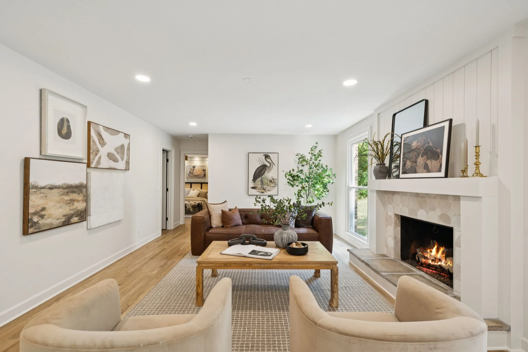Living room with white walls, hardwood floors, a fireplace, a brown leather sofa with pillows, a coffee table, framed artwork, and large windows with natural light.