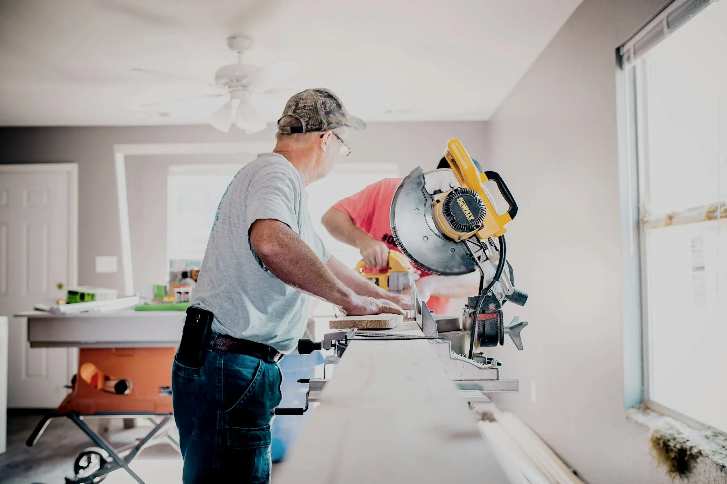 An elderly man wearing a camouflage baseball cap and glasses using a miter saw to cut wood in a bright, modern room under construction or renovation.