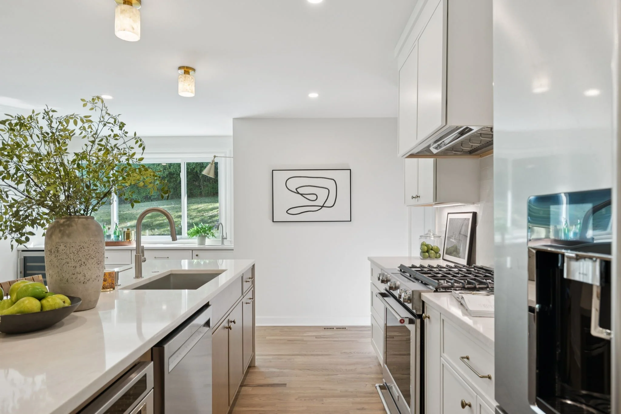 Modern kitchen with white cabinets and countertops, a large plant in a textured beige vase, a bowl of green apples, and a window showing greenery outside.