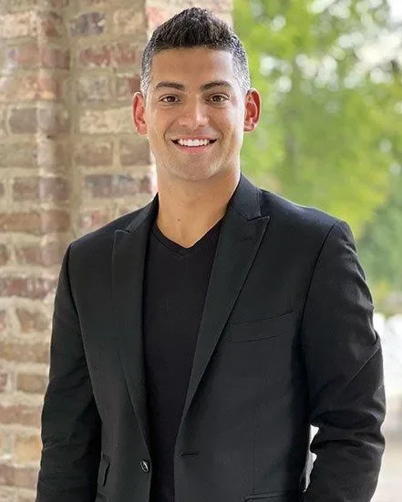 Young man smiling, wearing a black blazer and shirt, standing outdoors near a brick wall and green trees.