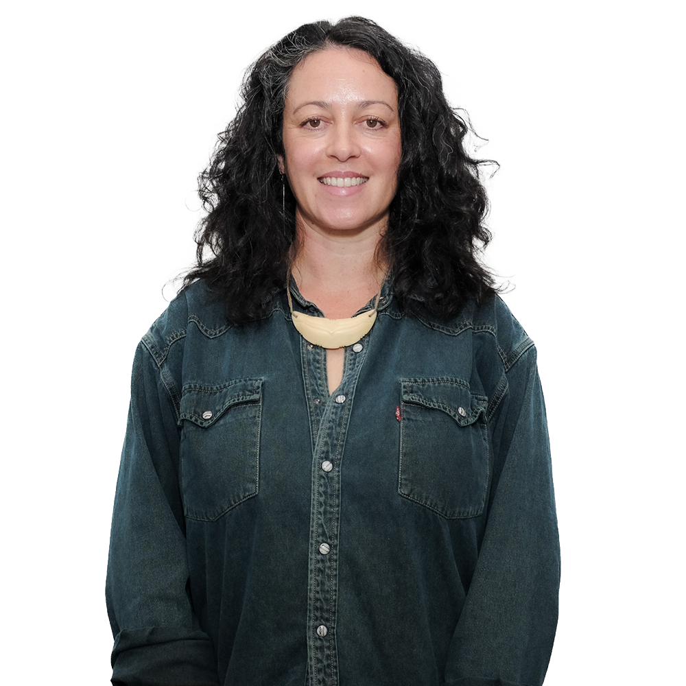 A woman with curly dark hair smiling, wearing a dark denim shirt and a cream-colored necklace.