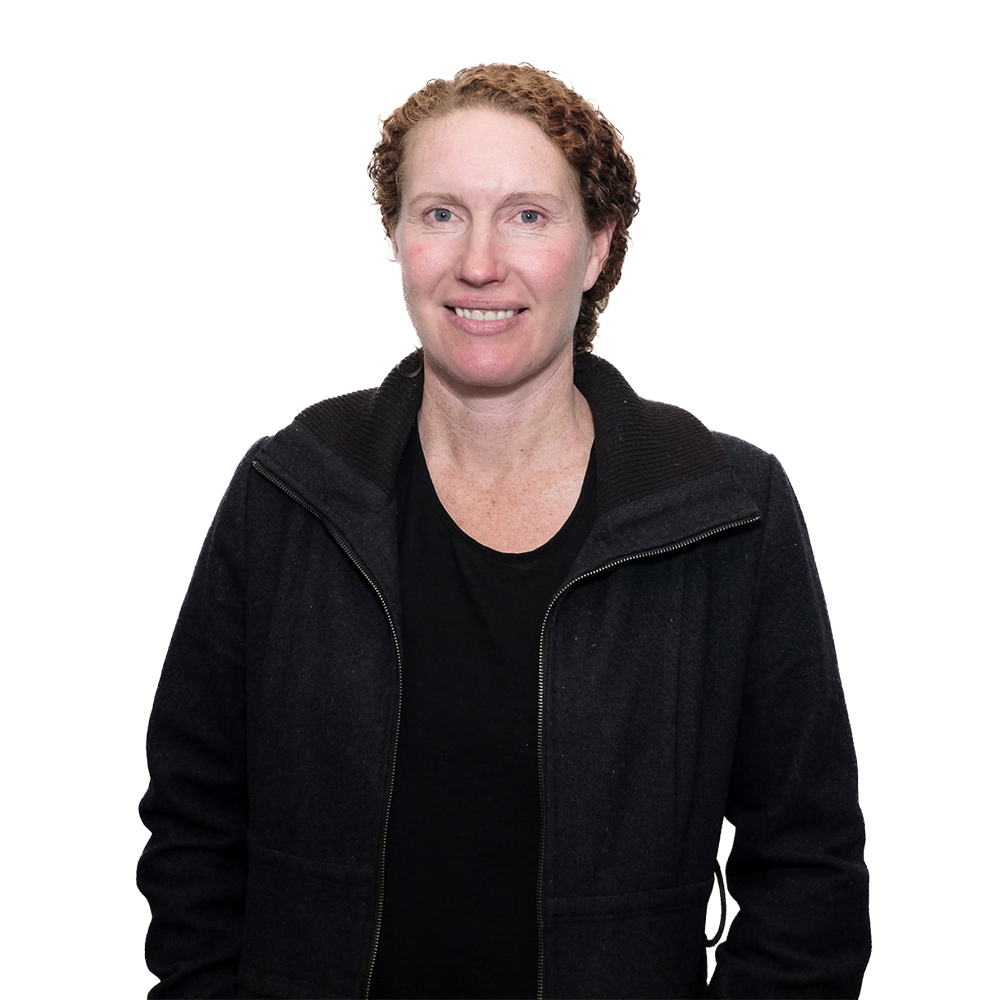 A woman with curly red hair smiling, wearing a black jacket and black shirt, standing against a plain background.