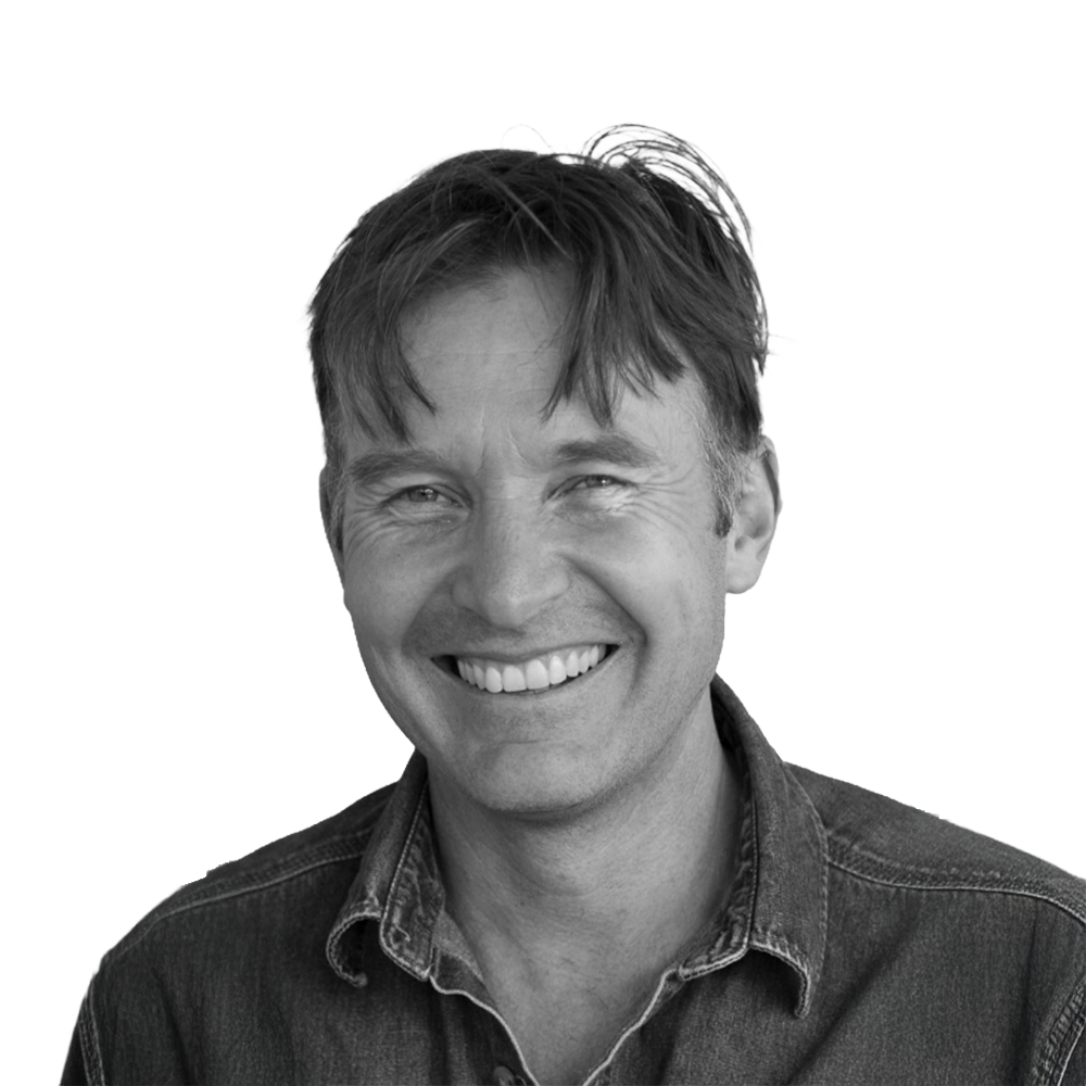 Black and white photo of a smiling man with medium-length hair, wearing a denim shirt.