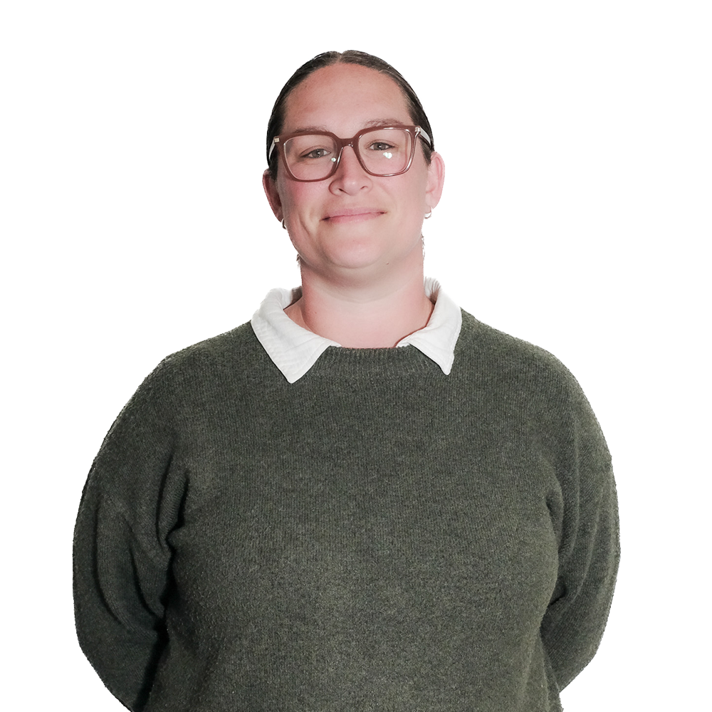 A woman with glasses, wearing a white collared shirt under a dark green sweater, smiling slightly, with her hands behind her back.