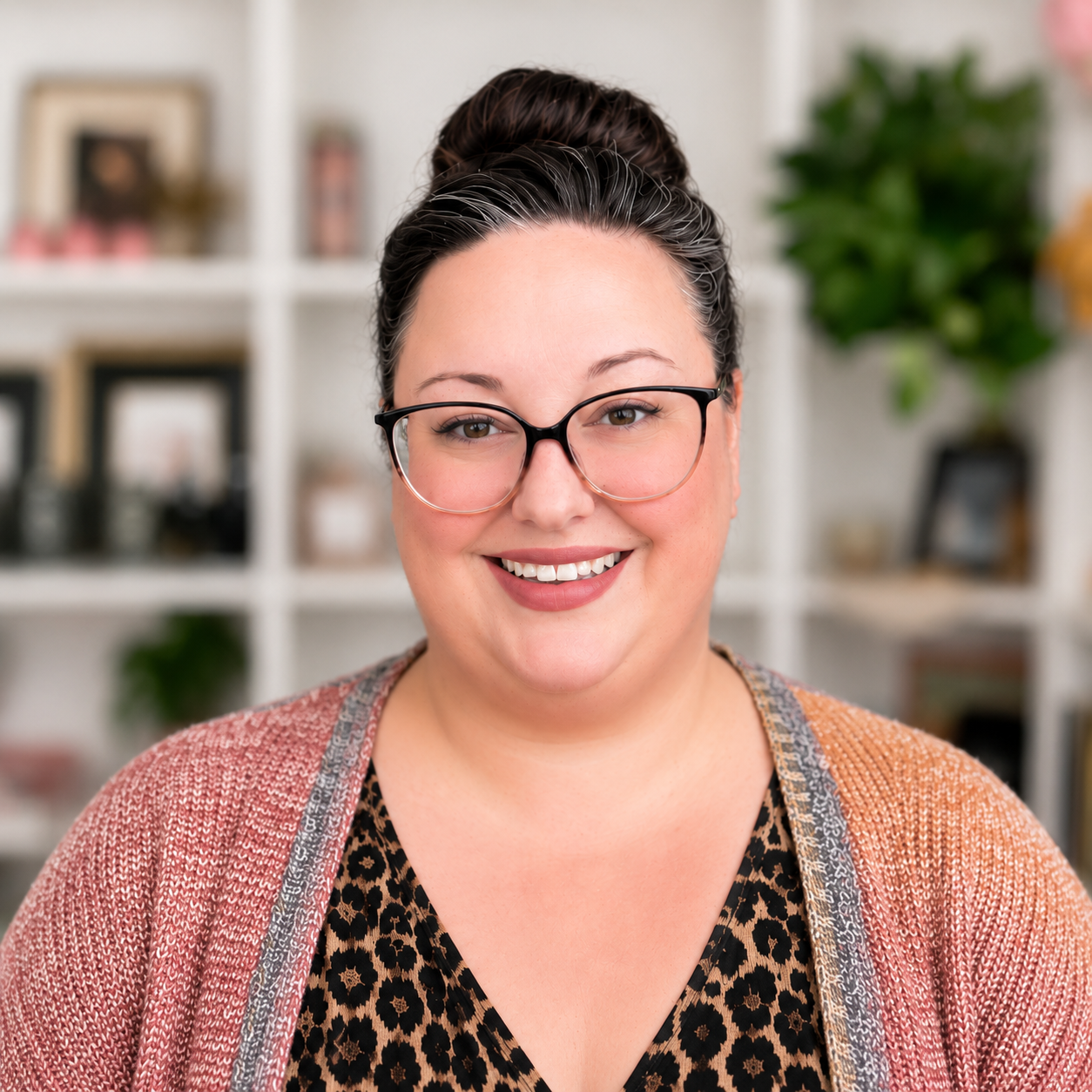 Smiling woman with glasses, dark hair in a bun, wearing a leopard print top and a pink and gray cardigan, in a cozy home setting with books and plants in the background.