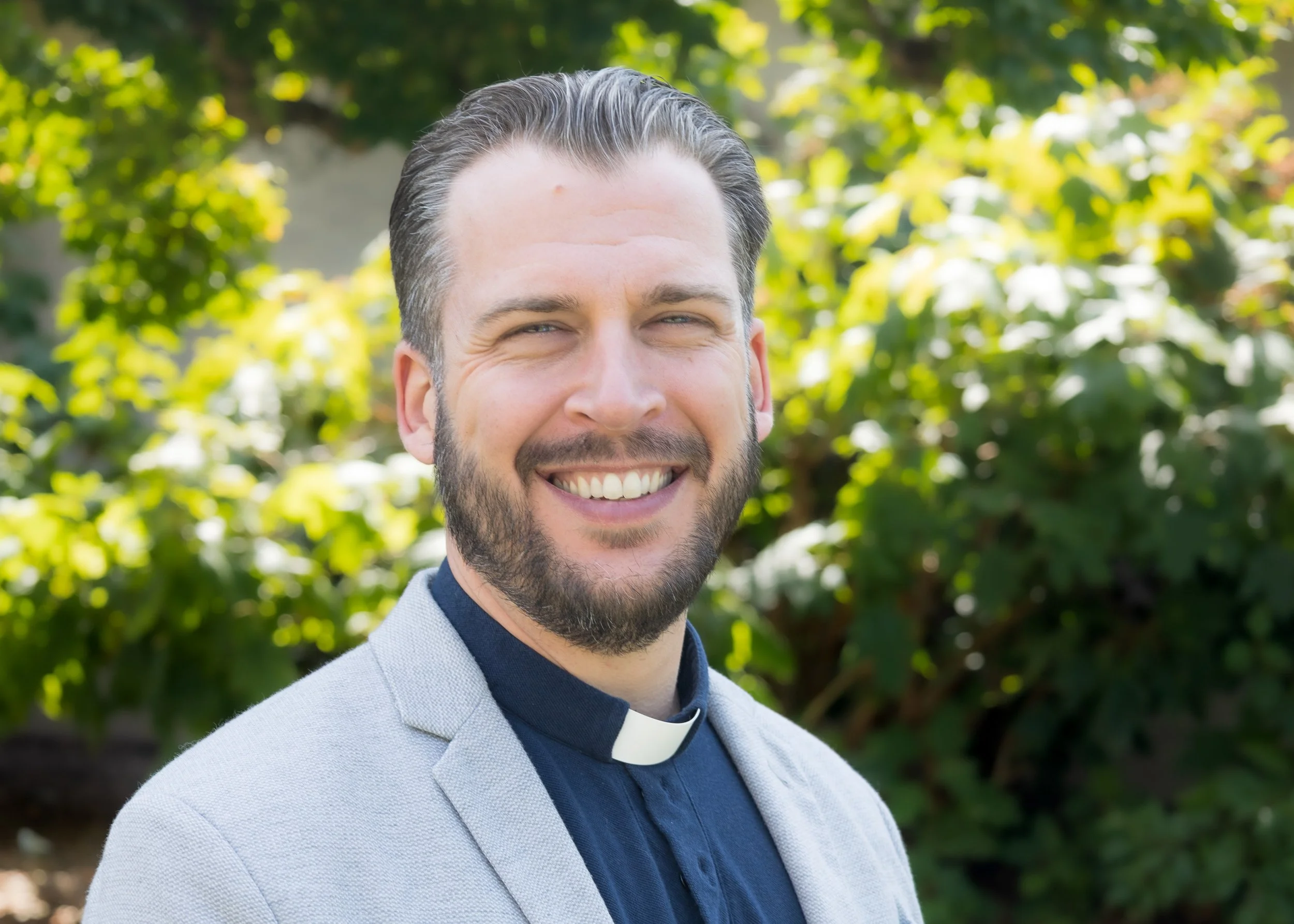 A smiling man with a beard and slicked-back hair, wearing clerical clothing with a gray blazer and a clerical collar, standing outdoors in front of green foliage.