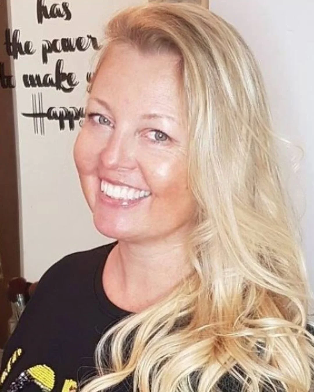 A smiling woman with long, wavy blonde hair, wearing a black shirt, standing in front of a whiteboard with black writing. The visible text on the whiteboard reads 'has the power to make'.