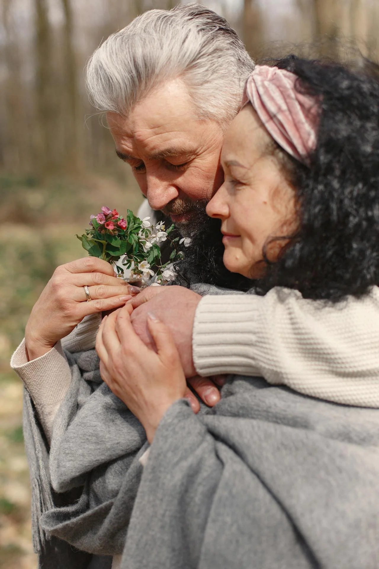 An older man and woman embrace outdoors, with the man holding a small bouquet of flowers, both smiling with eyes closed.