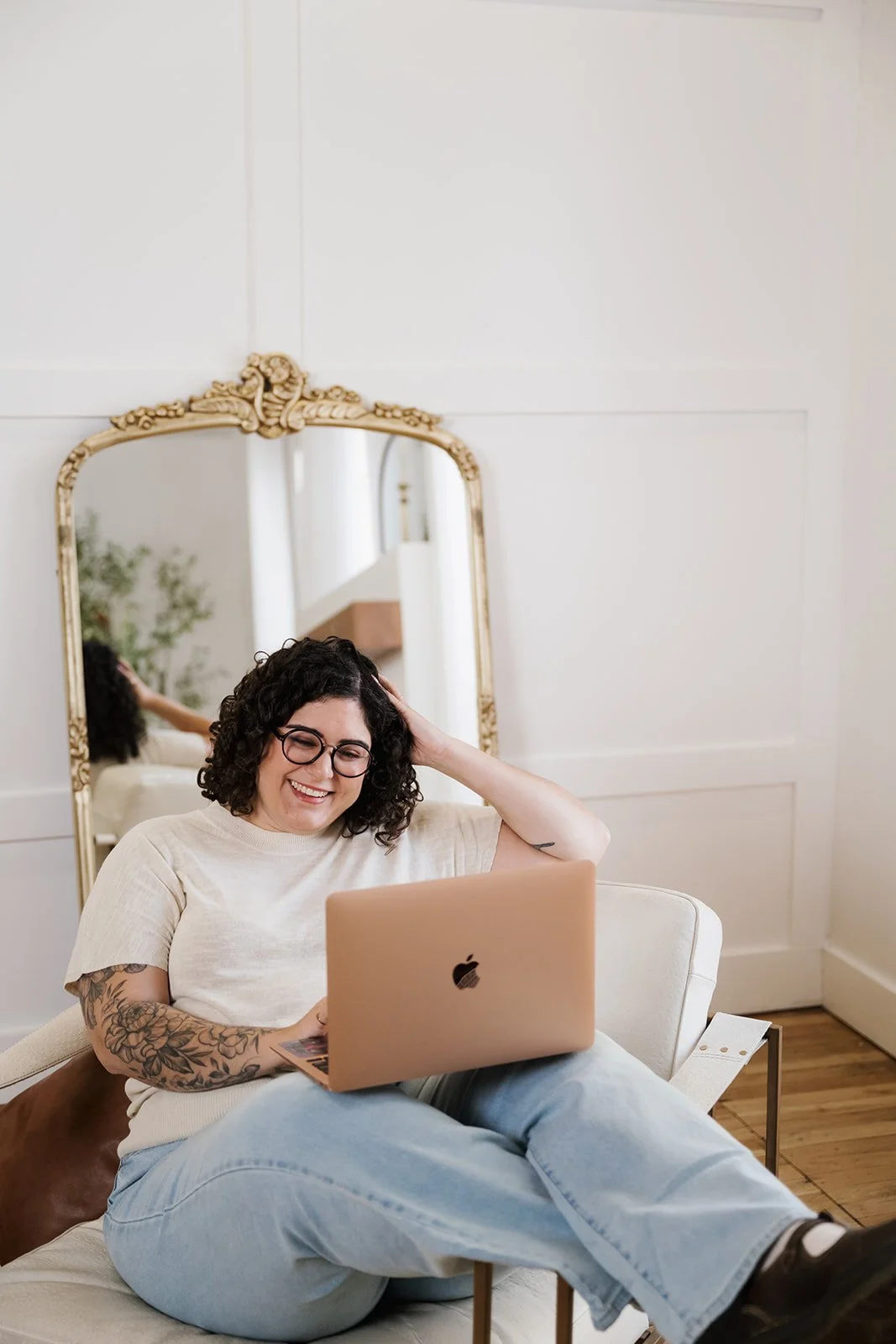 A woman with glasses and tattoos, sitting on a couch, smiling while using a pink laptop. Behind her, there is a large ornate mirror reflecting her arm and part of the room, with white walls and wood flooring.