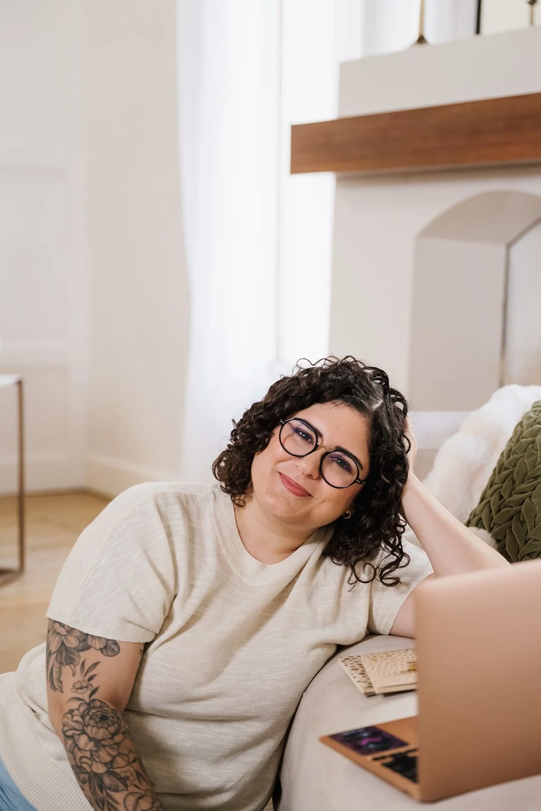 Woman with curly hair, glasses, and tattoos lying on a couch, smiling at the camera, with books and a laptop nearby in a bright room.
