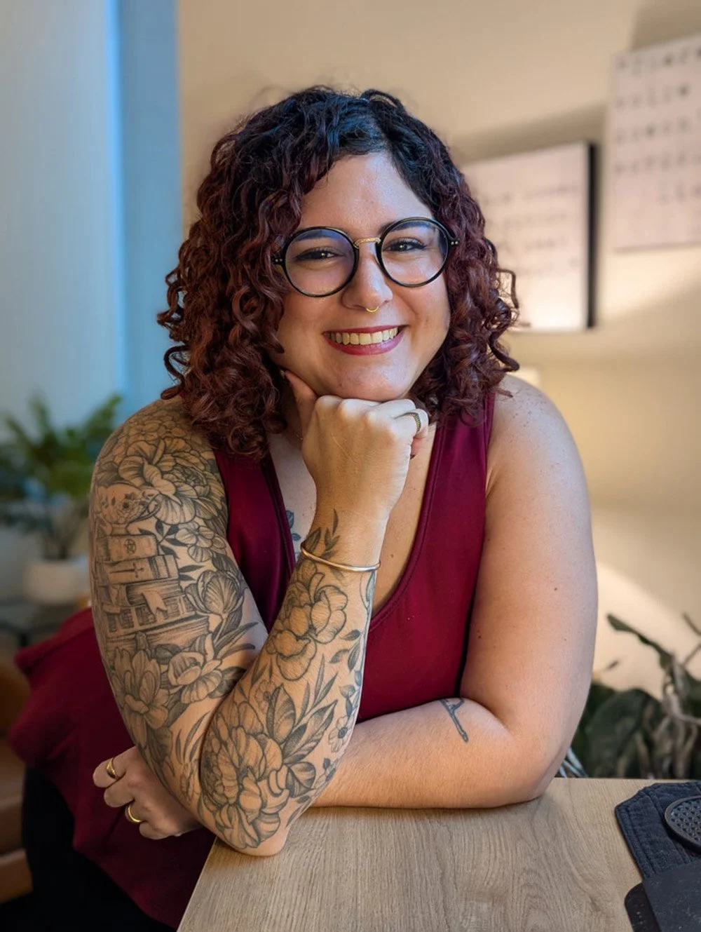 A smiling woman with curly dark red hair and glasses, wearing a sleeveless maroon top and jewelry, sitting at a table with her chin resting on her hand in a brightly lit room.