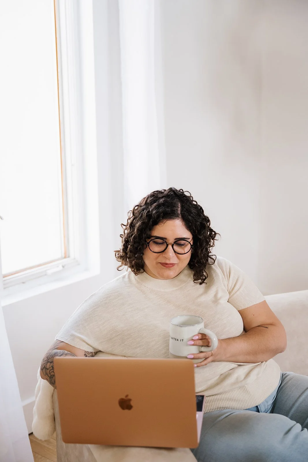 A woman with curly hair and glasses sitting on a beige couch, holding a white mug, working on a laptop in a bright, minimalistic room with white walls and a window.