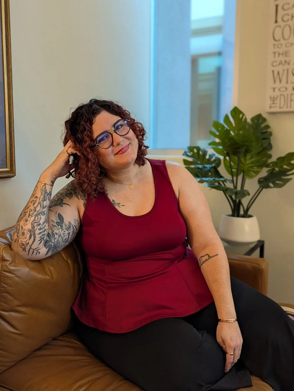 A woman with curly dark hair and glasses sitting on a brown leather couch, leaning her head on her hand, wearing a red sleeveless top and black pants, in a well-lit room with a plant and artwork on the wall.