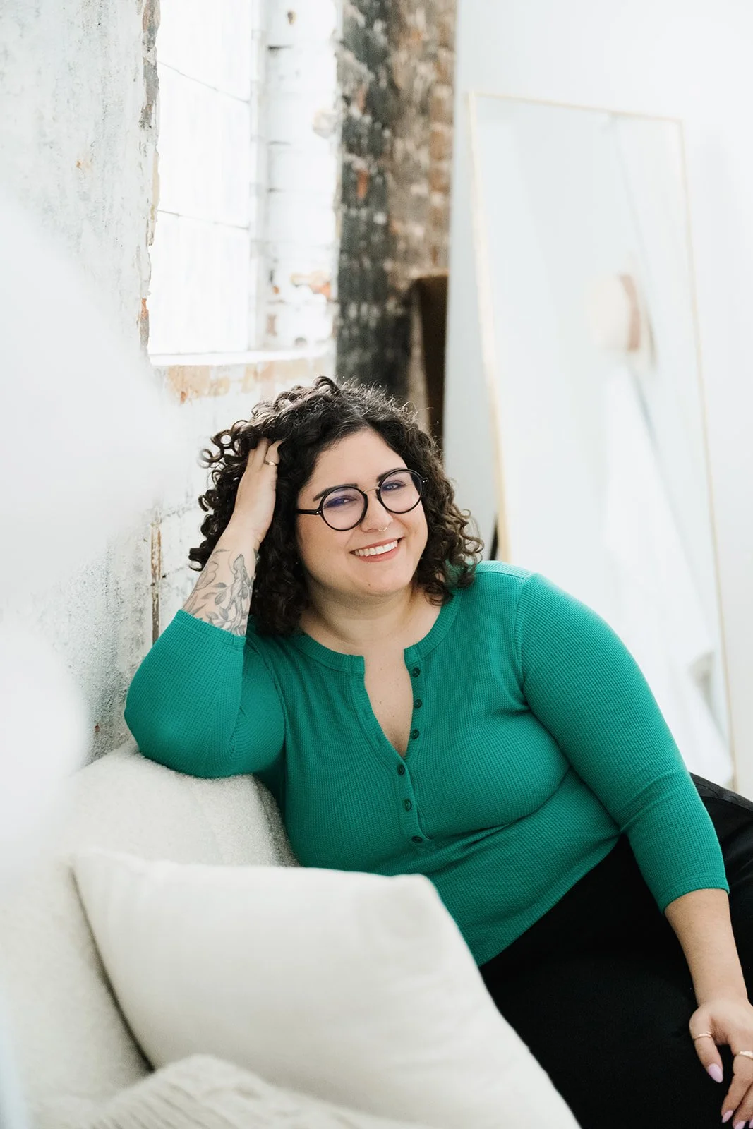 A woman with glasses and curly hair wearing a green top, smiling and relaxing on a white couch in a bright, industrial-style space.