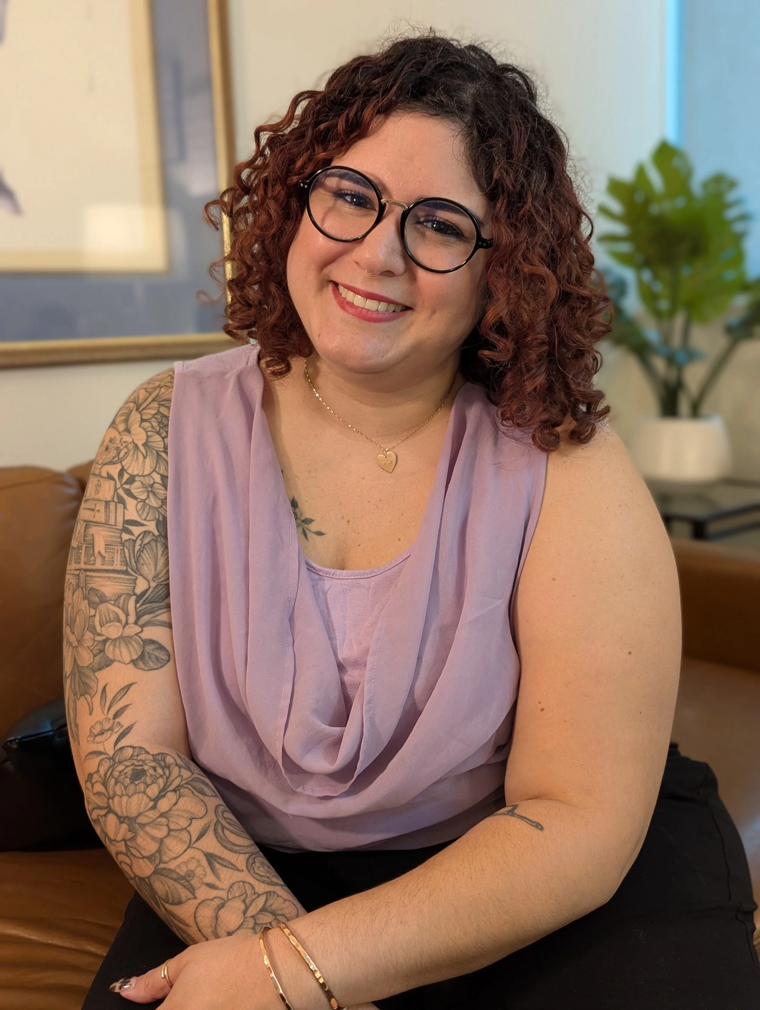 A woman with curly red hair, wearing glasses, a sleeveless lavender top, and a gold heart necklace, sitting on a couch in a room with a potted plant and framed pictures in the background.