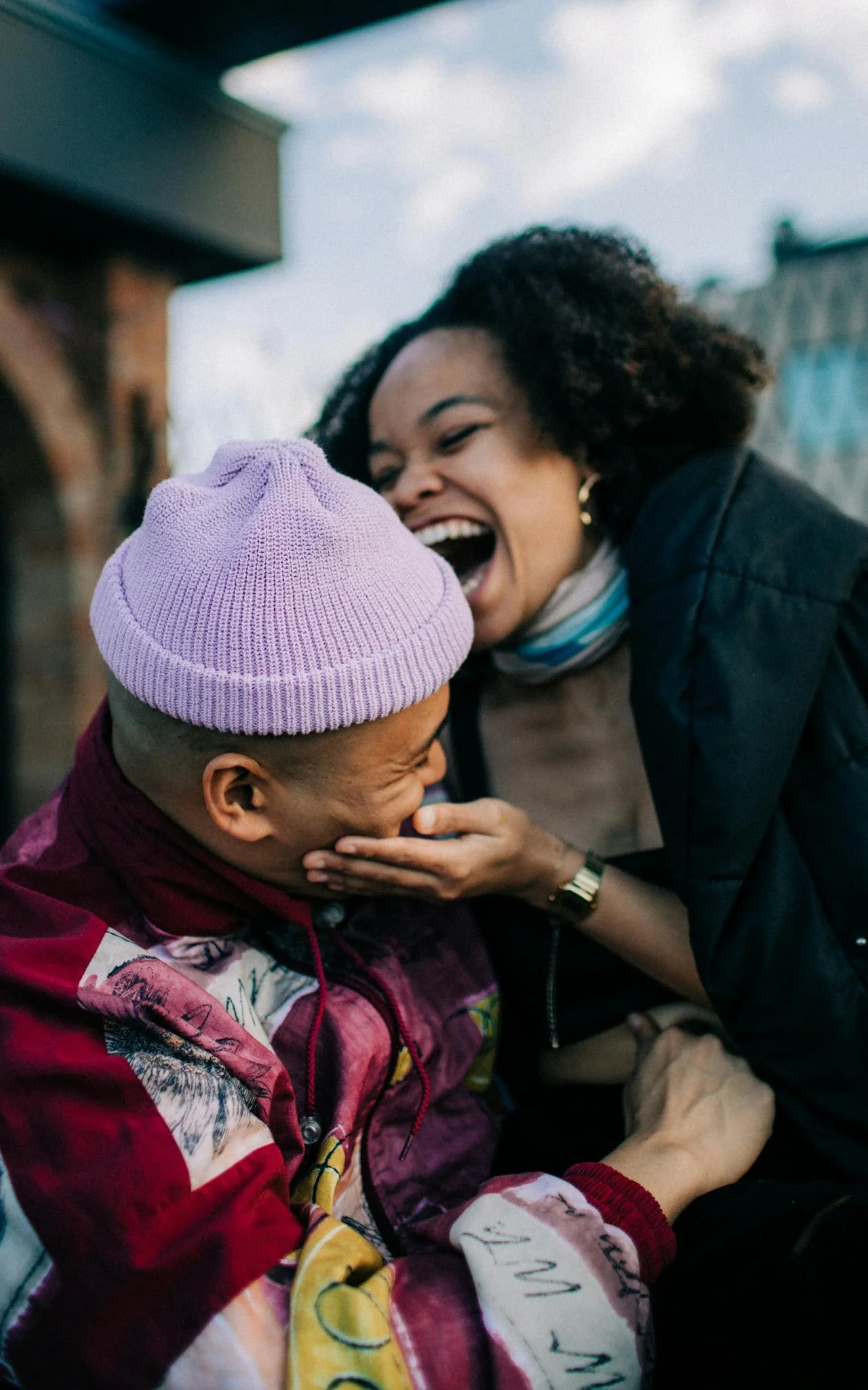 Two women sharing a joyful moment outdoors, one laughing and causing the other to cover her face, against a cloudy sky and building in the background.