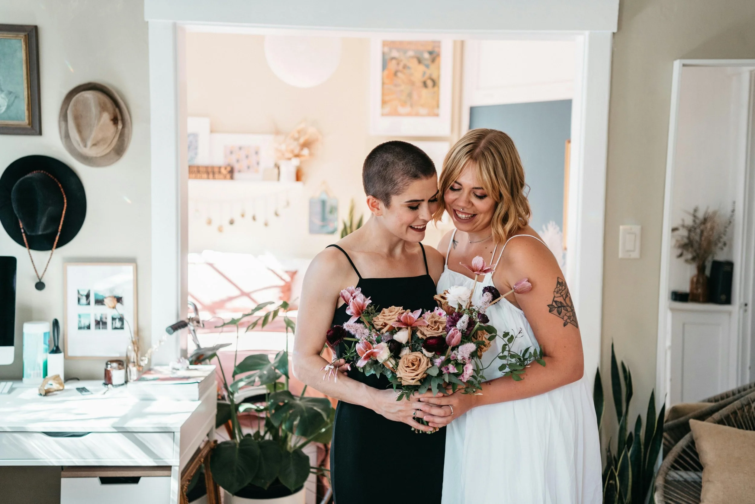 Two women smiling and hugging, one holding a colorful bouquet of flowers, inside a cozy, decorated home.