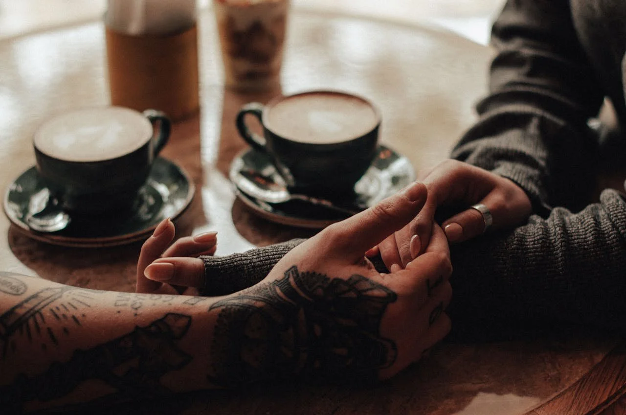 Two people holding hands on a wooden table with two cups of coffee, two glasses of iced coffee, and a glass of iced tea.