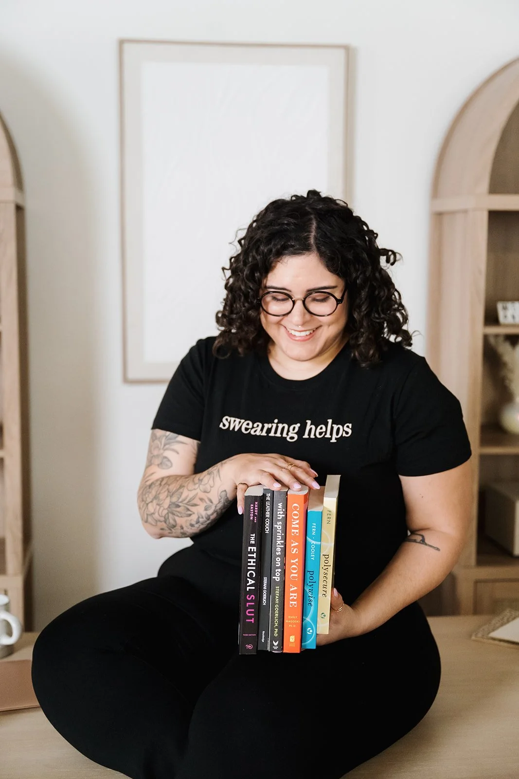 Woman with curly hair and glasses smiling while holding a stack of five books, sitting cross-legged on a wooden floor in a cozy room.