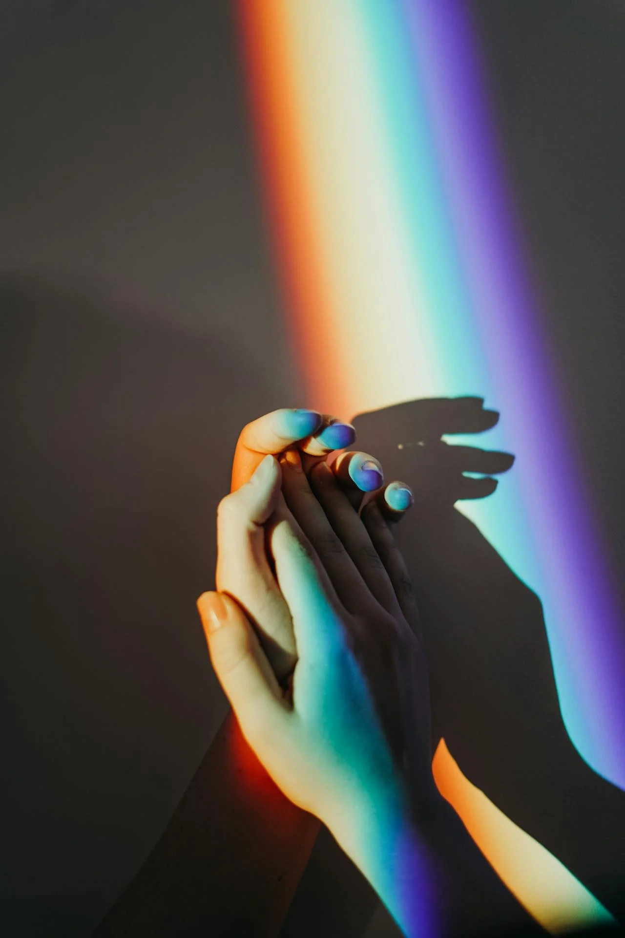 Two hands with intertwined fingers casting a rainbow-colored shadow on a dark background