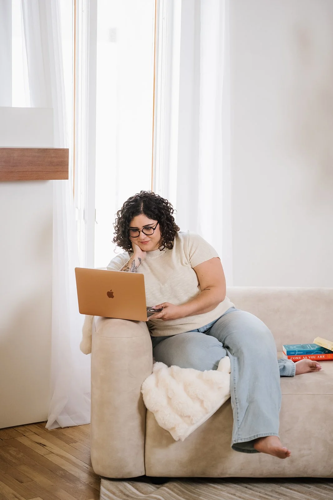 A woman with curly dark hair and glasses is sitting on a beige couch, working on a gold MacBook. She is wearing a cream shirt and light blue jeans, with her feet resting on the couch. There are books on the side of the couch, and a blanket is draped over her legs. The room has white walls and large windows with white curtains, creating a bright and cozy atmosphere.
