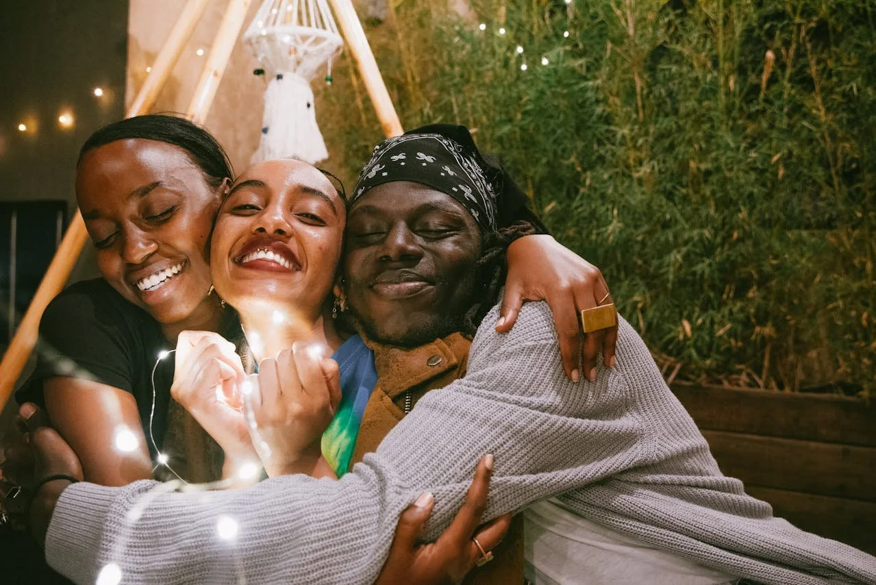 Three people hugging and smiling at the camera in a cozy outdoor setting with warm lighting and greenery.