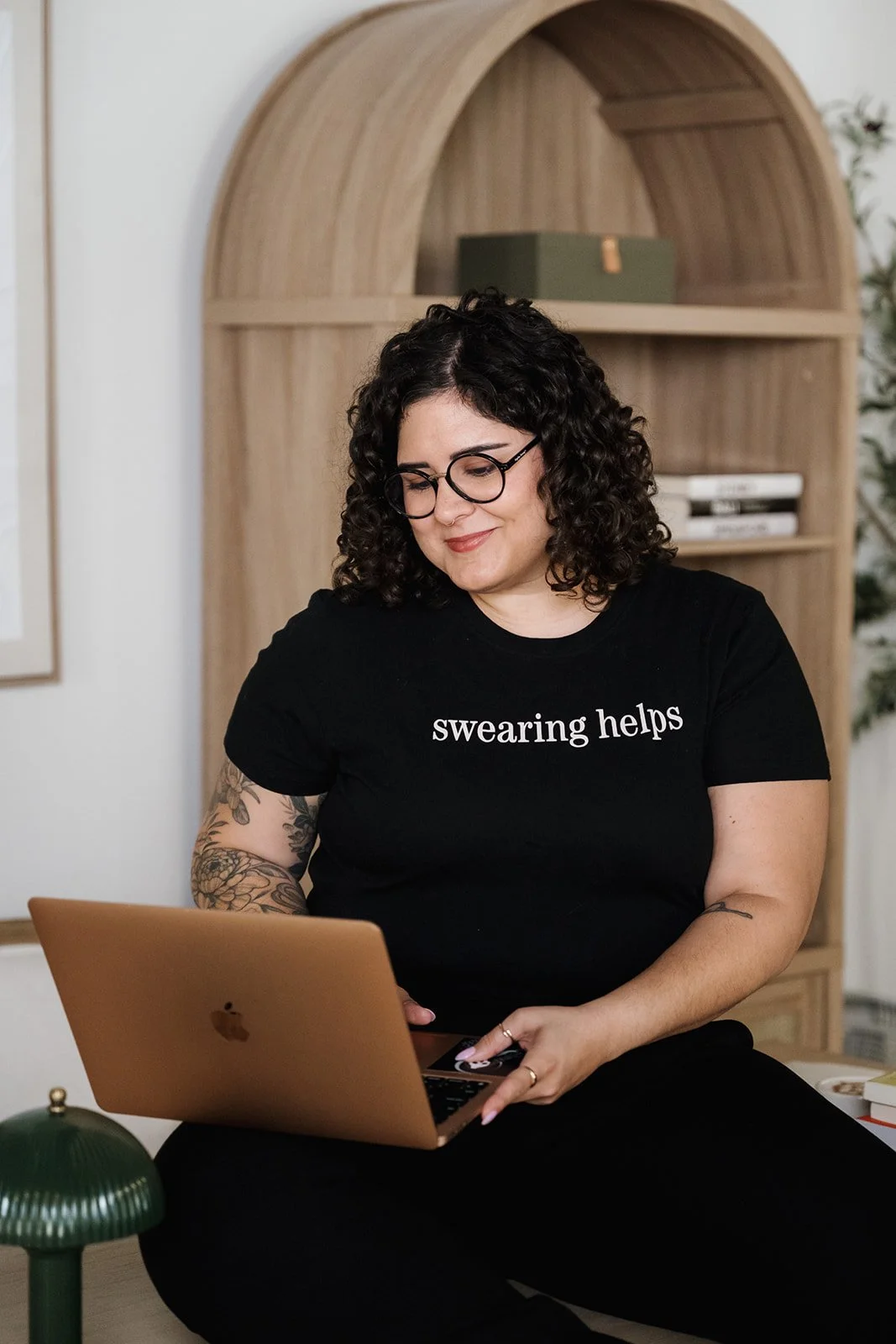 A woman with curly dark hair and glasses, wearing a black t-shirt that says 'swearing helps,' sitting on the floor and working on a laptop with a wooden bookshelf in the background.