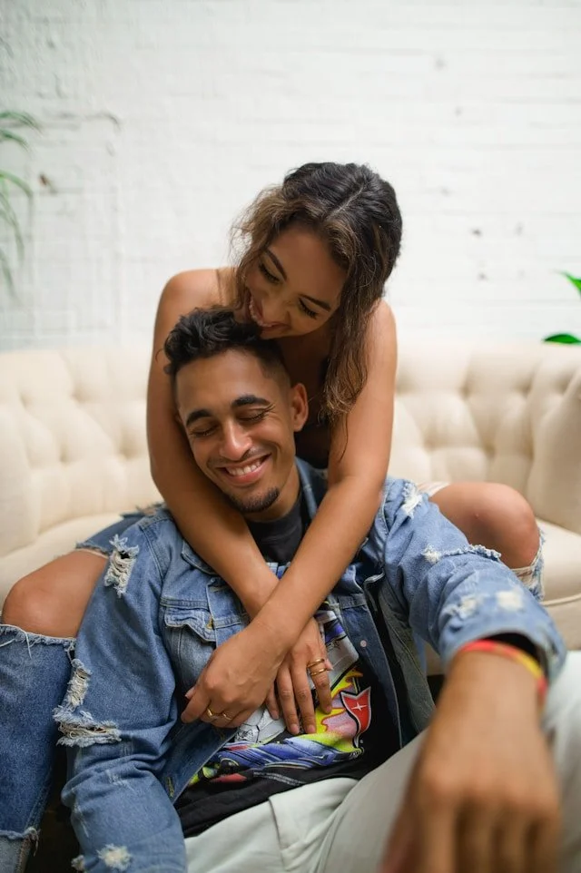 A woman and a man sitting on a cream-colored couch, smiling and embracing each other affectionately indoors with a white brick wall in the background.