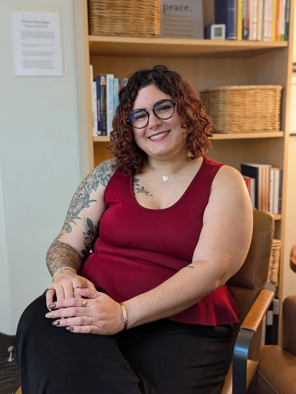 A woman with curly red hair and glasses, wearing a red sleeveless top, sitting in a wooden chair with a bookshelf behind her, smiling at the camera.