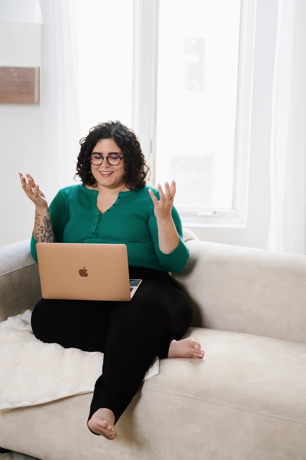 Woman sitting on a beige couch with a MacBook laptop on her lap, smiling and gesturing with her hands, in a bright room with large windows and white curtains.