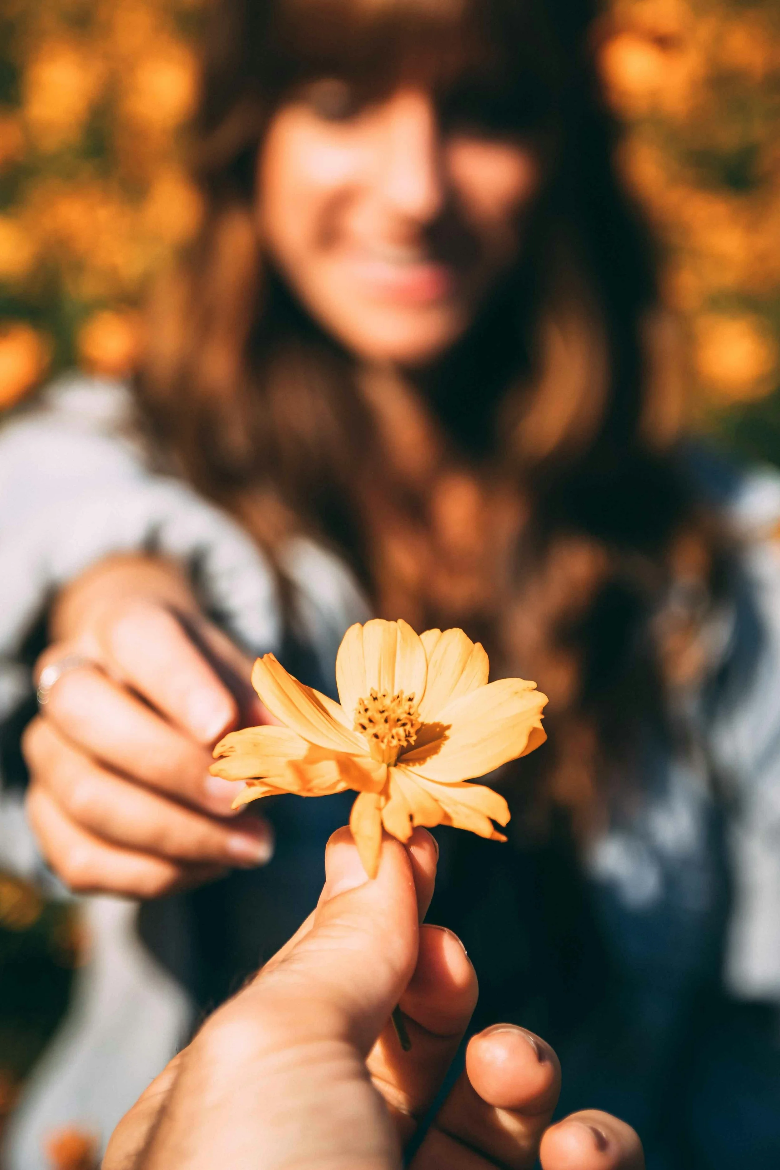 A woman with dark hair is smiling as she receives a light orange flower from a person in the foreground, with a background of autumn leaves.