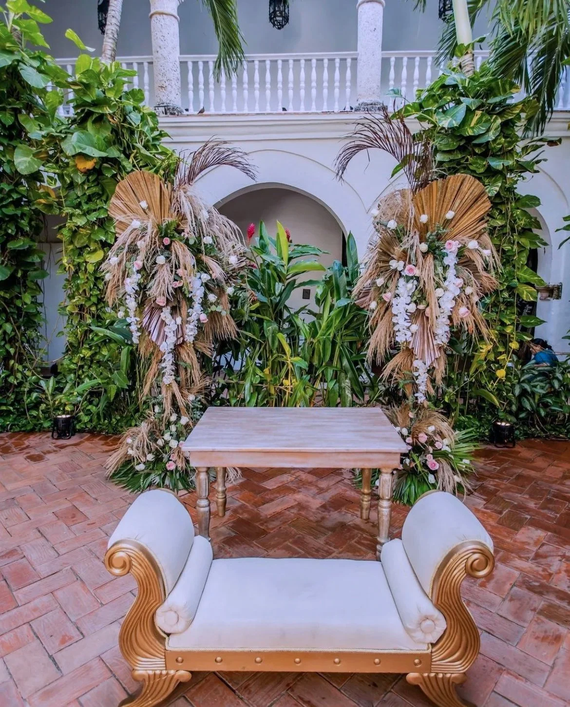 Decorative outdoor wedding altar with tropical foliage, dried palm leaves, and flowers, set in a courtyard with tiled floor and white architectural elements.