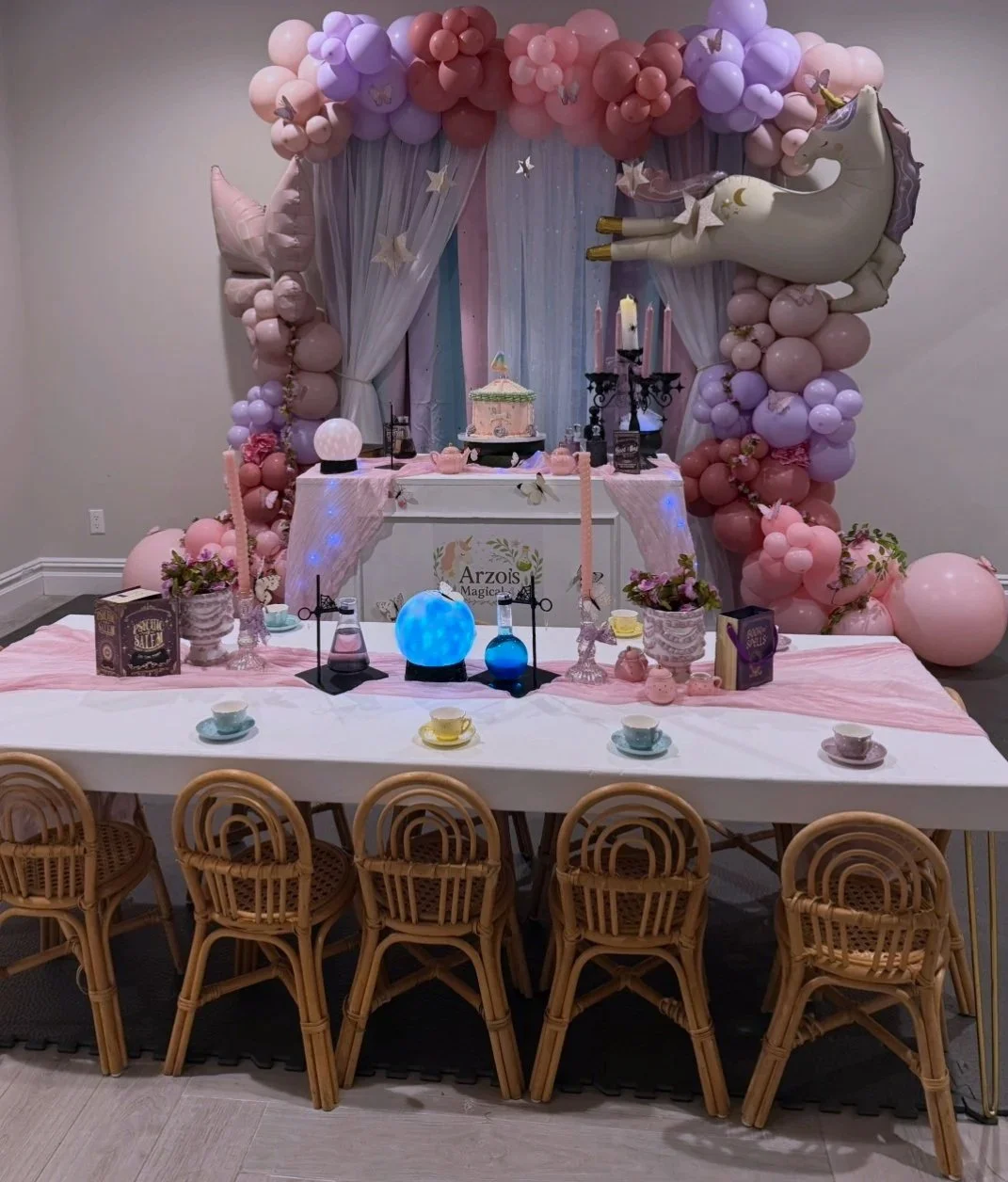Decorative unicorn-themed birthday party setup with pink and purple balloons, a unicorn balloon, a cake on a table, candles, and tea cups on the table.