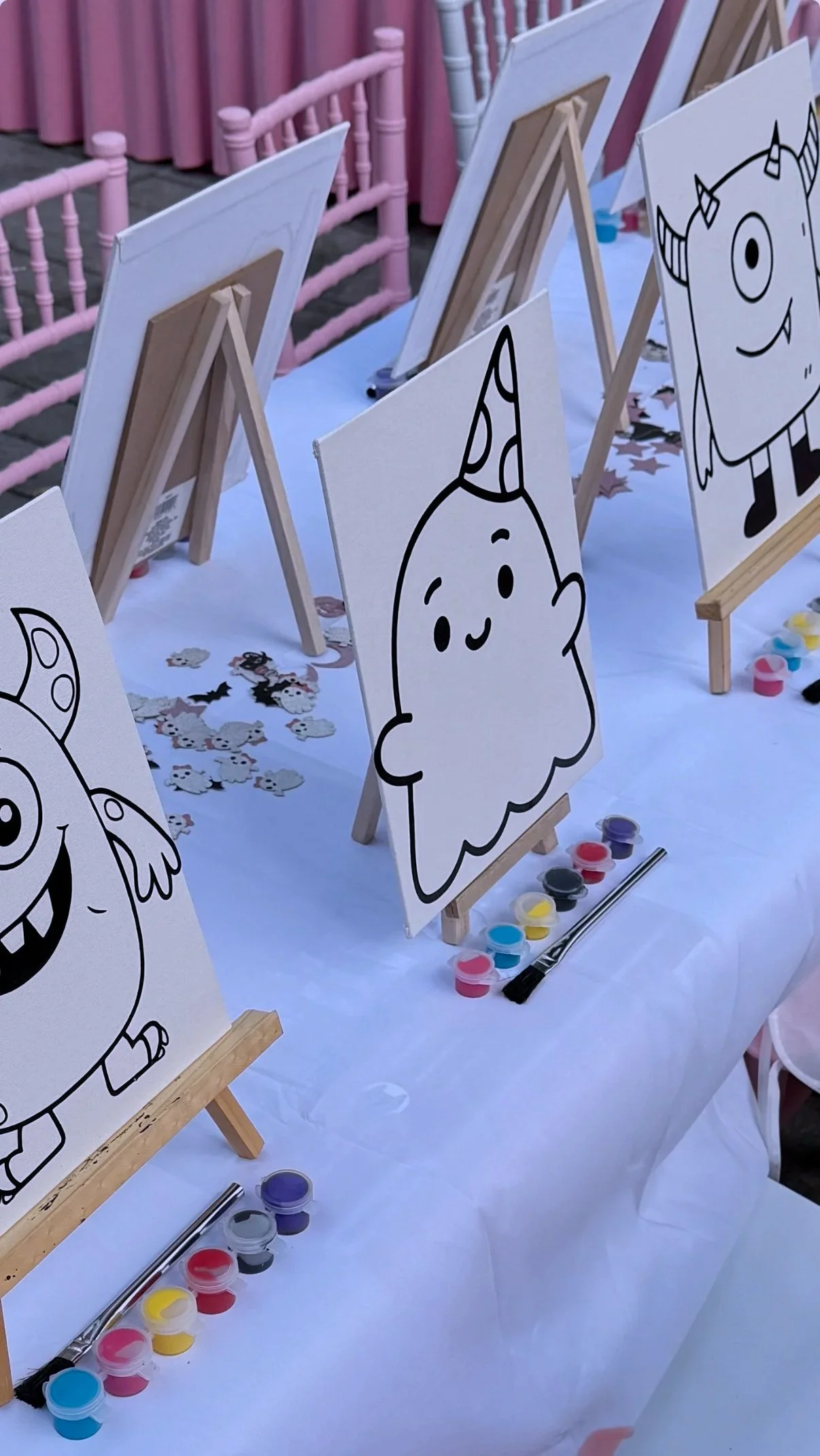 Tables with canvas boards of cartoon character outlines, paint pots, and paintbrushes for a coloring activity, with paper confetti and decorations in the background.