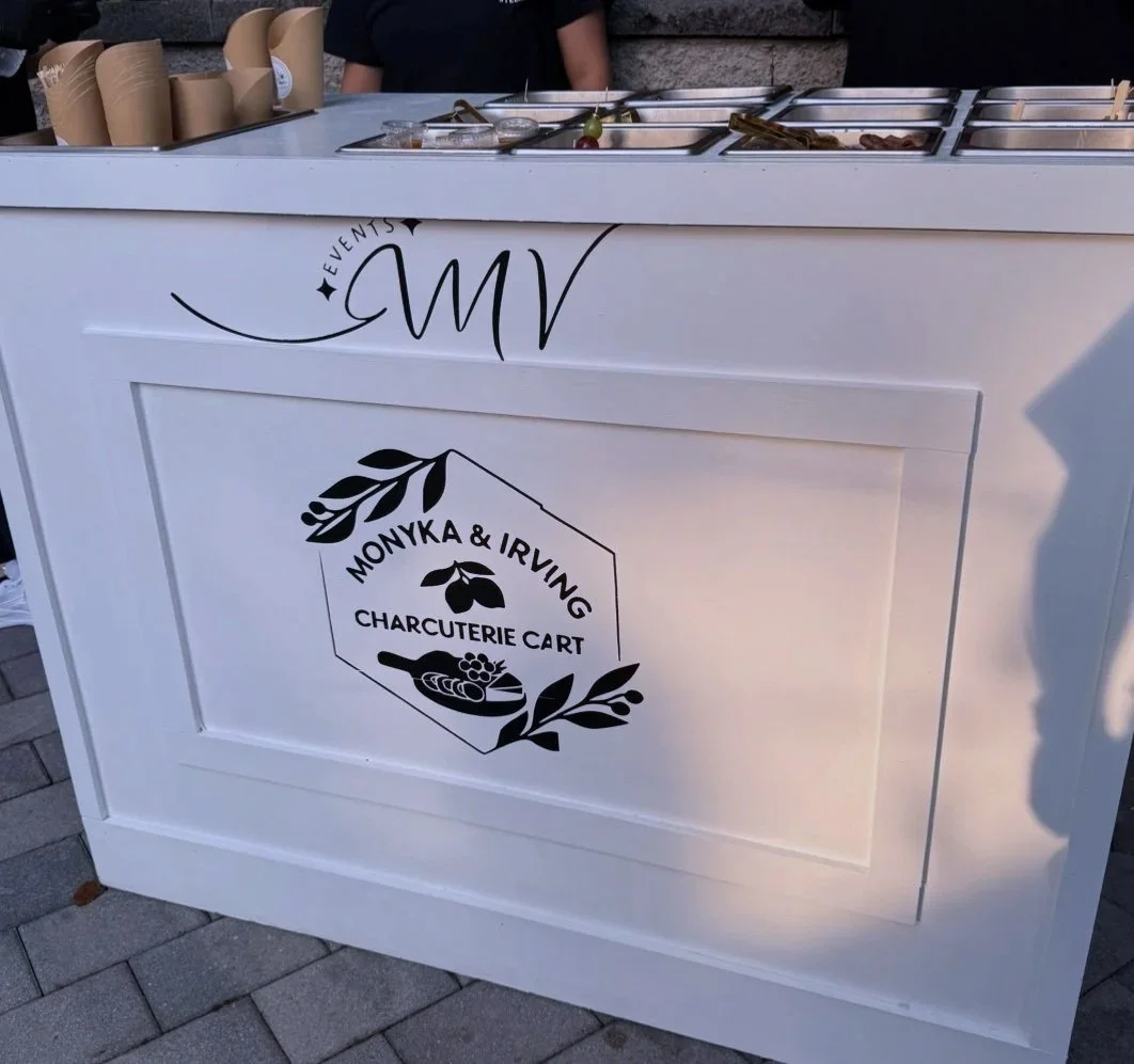 A white food cart with a sign that reads "MVM Events" on top and "Monyka & Irving Charcuterie Cart" on the front, along with a decorative illustration of a leaf and a platter of meats and cheese.