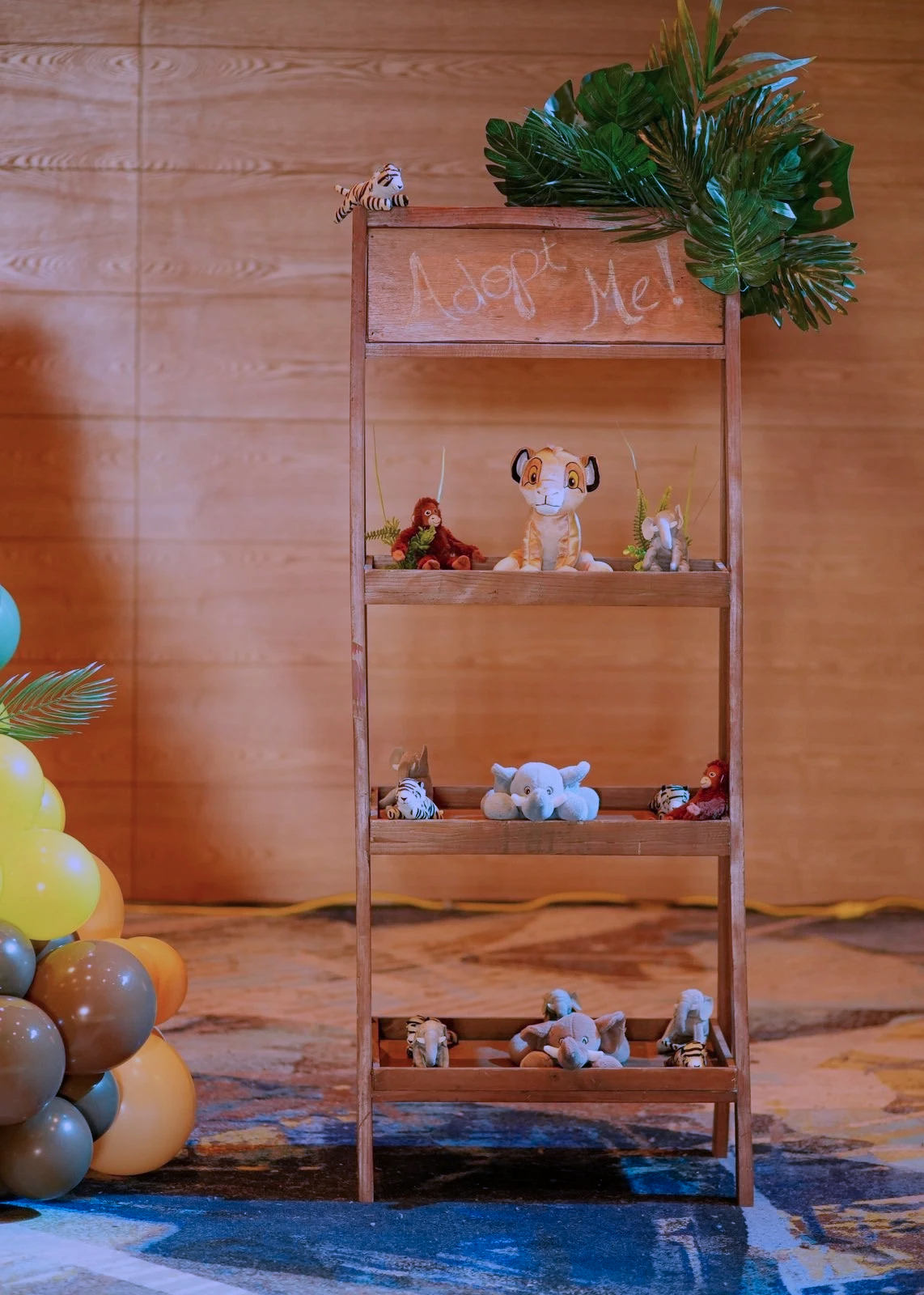 Wooden shelving unit with stuffed animal toys including zebras, elephants, lions, and tigers, against a wood-paneled wall. A small sign on the top shelf reads 'Adopt Me!'.