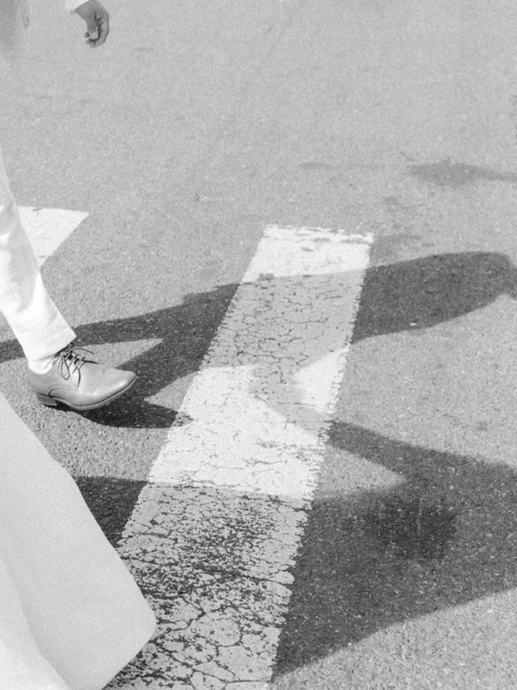 Foot of a person in white shoes and light-colored pants standing on a cracked pedestrian crosswalk on asphalt, with their shadow and part of their leg visible.