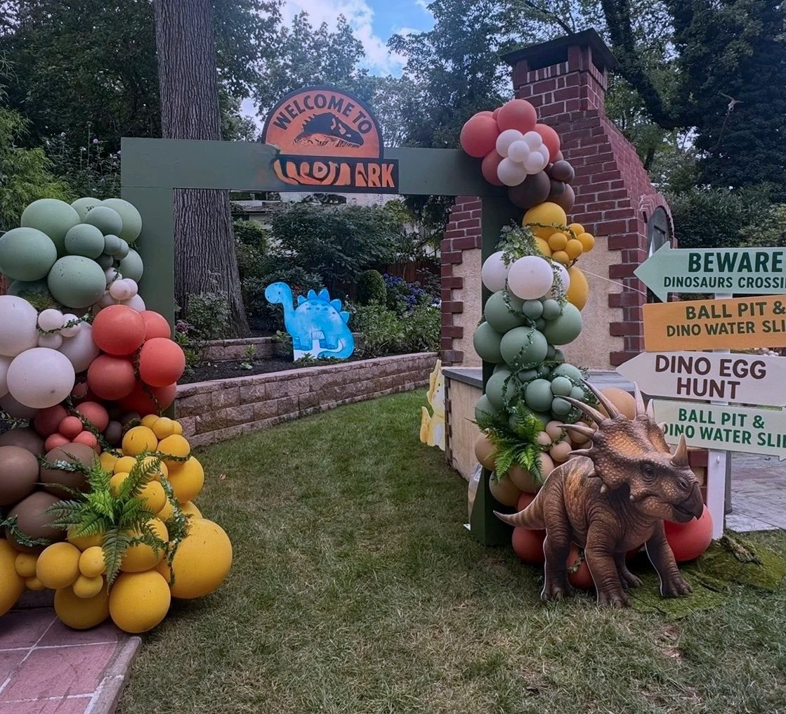 Decorated garden entrance with balloons, a dinosaur figure, and signs indicating Dino Crossing, Dino Water Slide, Dino Egg Hunt, and Ball Pit near a sign that reads "Welcome to Jurassic Park."