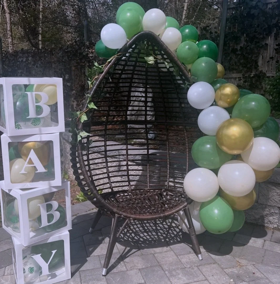 A decorative outdoor setup featuring a dark wicker egg-shaped chair surrounded by green, white, and gold balloons, with white cubes spelling out 'BABY' on the left.