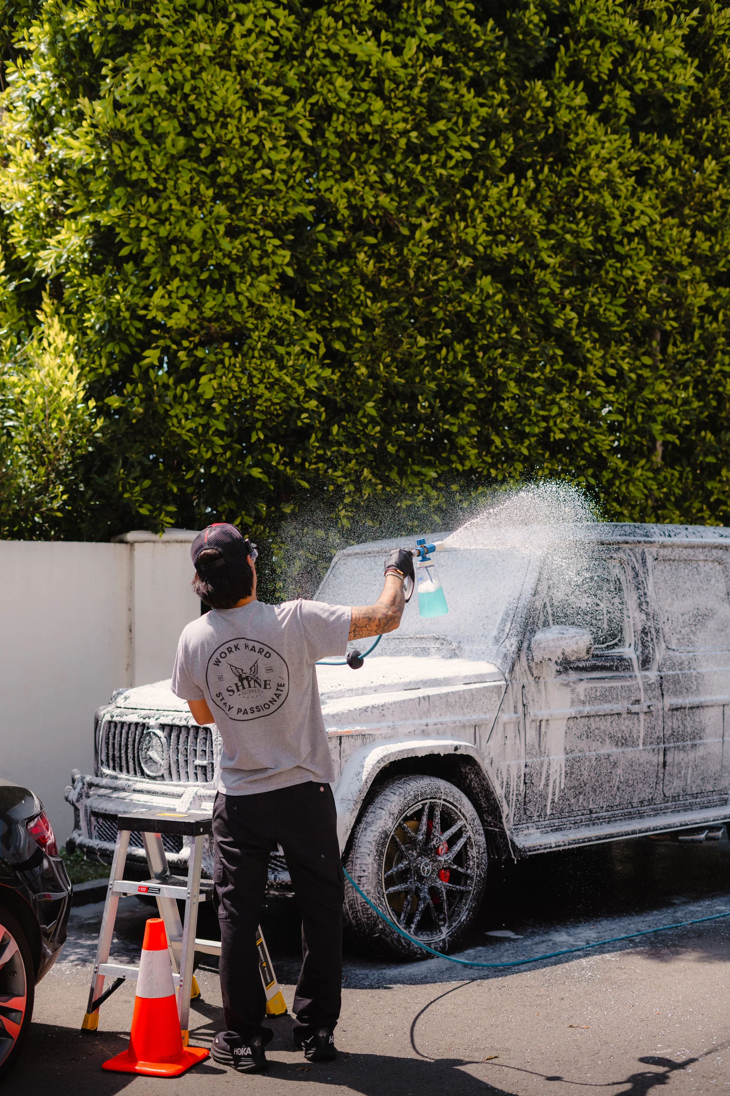 Person washing a white SUV with soap and water outside in a driveway, with a large green bush and a white wall in the background.