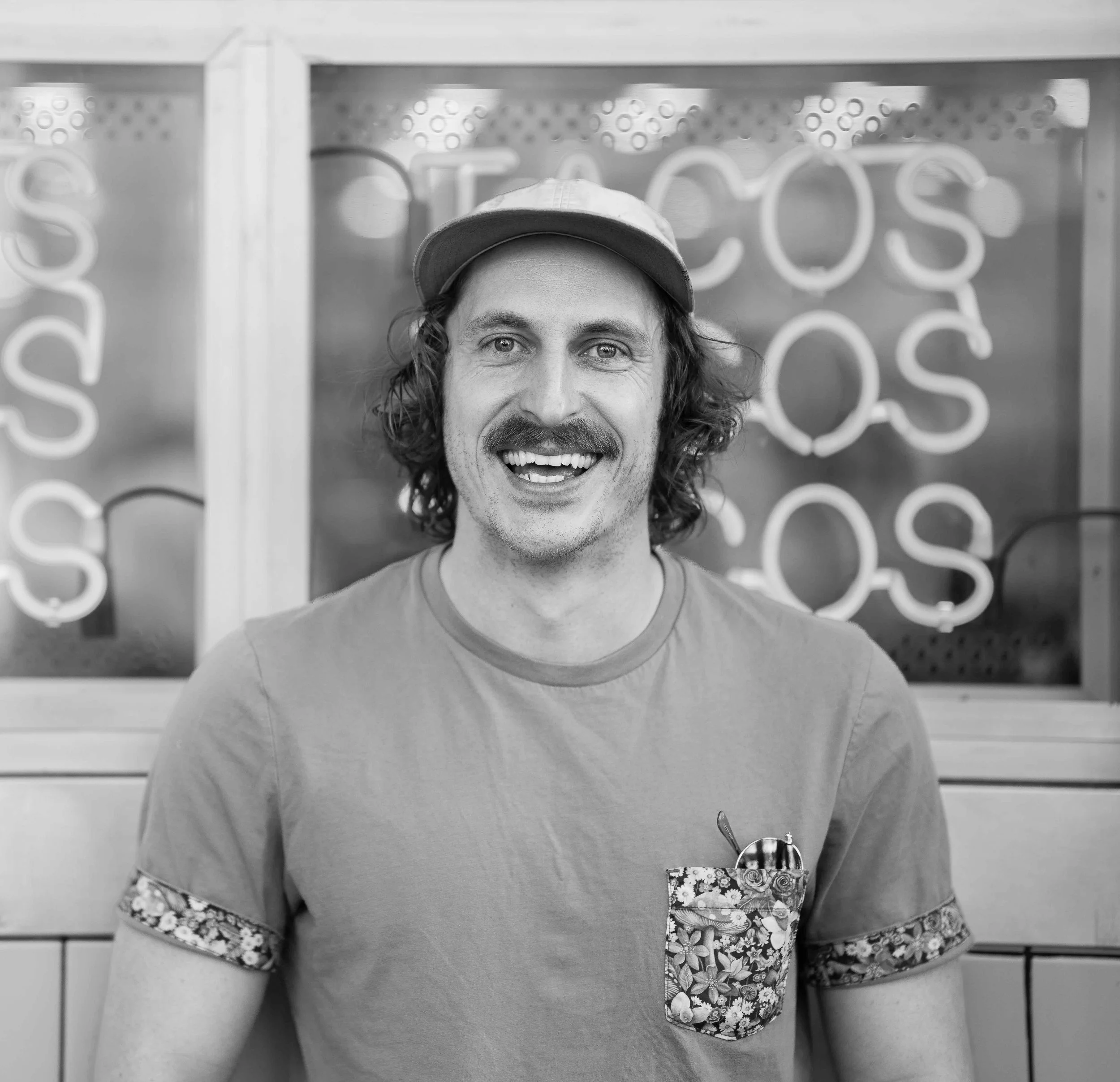 A smiling man with curly hair and a mustache, wearing a cap and a t-shirt with a floral pocket, standing in front of a neon sign background.