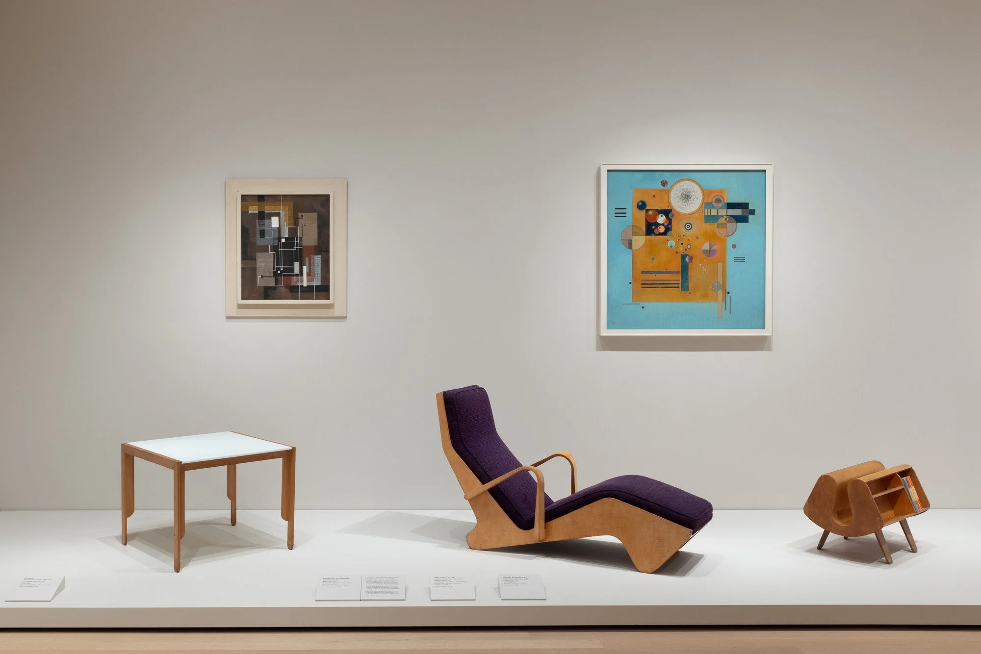 Display of modern furniture and artwork in an art gallery. Features a small table, a purple upholstered lounge chair, and a small bookshelf or storage unit, with two abstract paintings on the white wall behind.