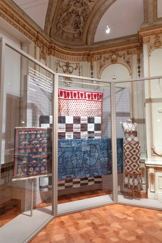 Display of various textile art pieces, including woven tapestries and embroidered textiles, in an ornate room with gold detailing on walls and ceiling.