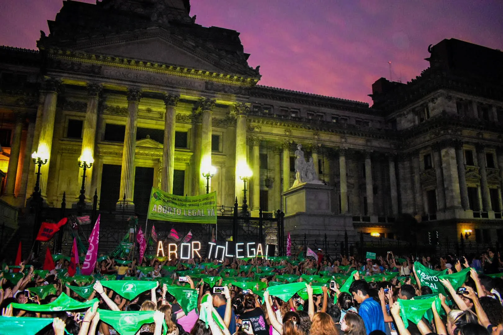 Pro-choice protest in front of a historic building at dusk, with people holding green flags and signs supporting legal abortion.