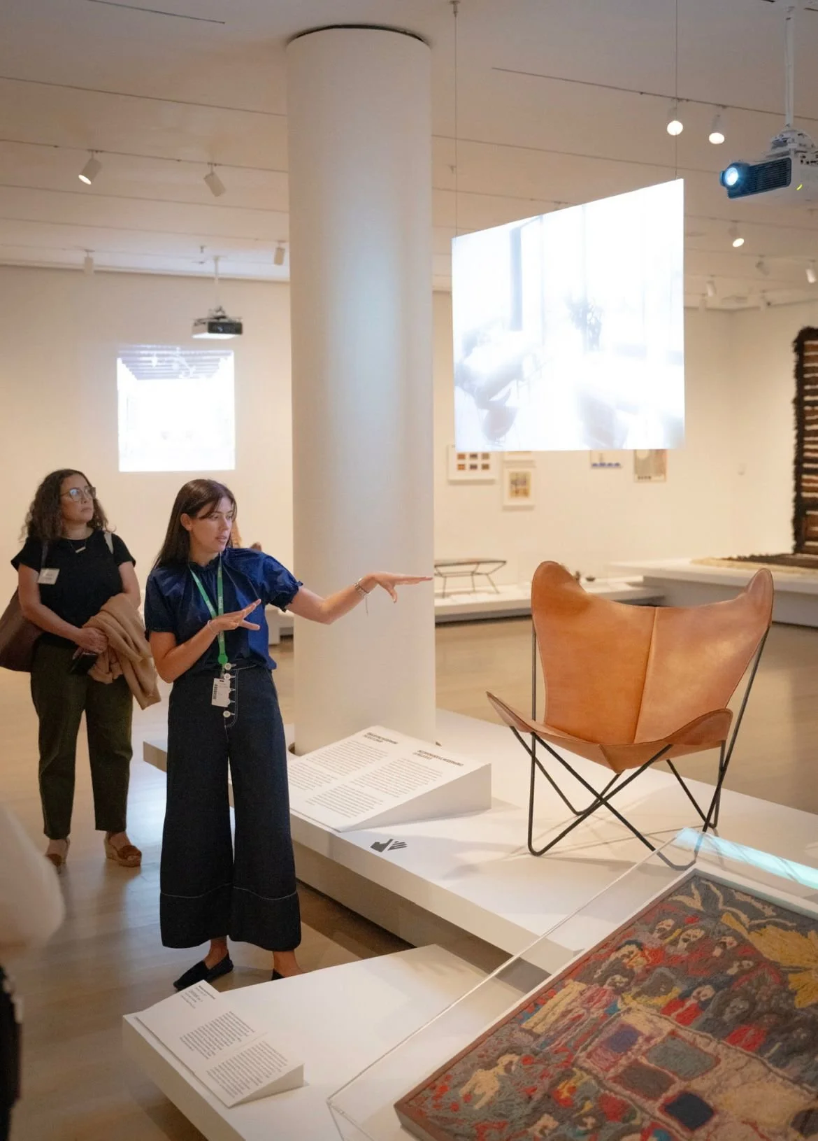 Two women in an art gallery, one gesturing towards a modern wooden chair displayed on a white platform, with informational plaques nearby. A large screen and artwork are visible in the background.