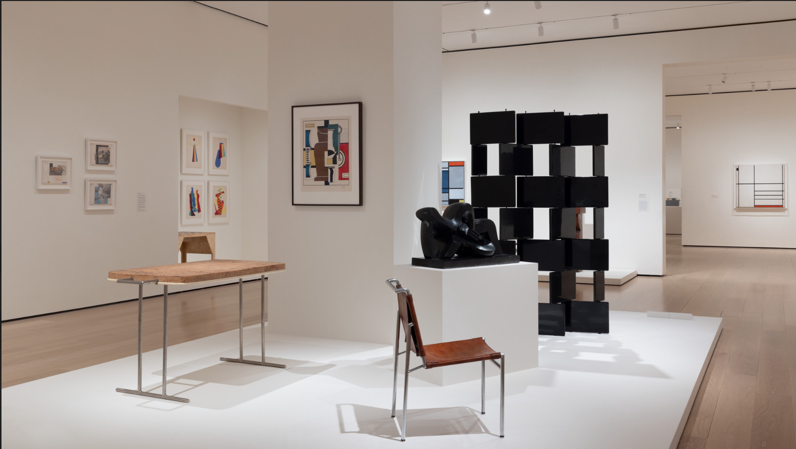 Interior of an art gallery displaying various modern sculptures and paintings on white walls, including a black modern sculpture on a white pedestal, a abstract right-angled black sculpture, and colorful abstract paintings.