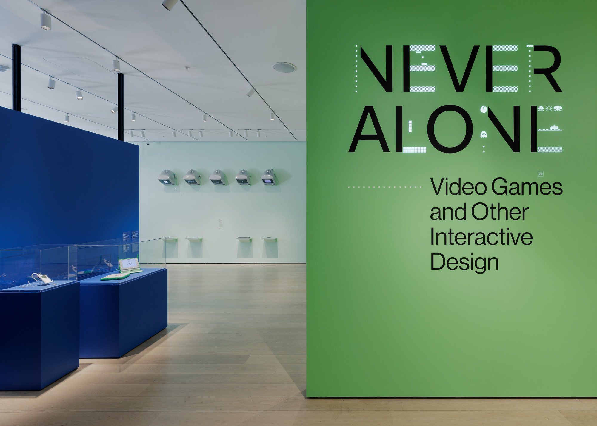 Exhibit space titled 'Never Alone' featuring a green wall with black text about video games and interactive design, with display cases and wall-mounted devices in a gallery setting.