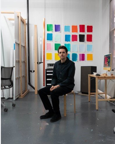 A man seated on a chair in an art studio with a colorful grid of paint swatches on the wall behind him, and a computer on a wooden desk to his right.